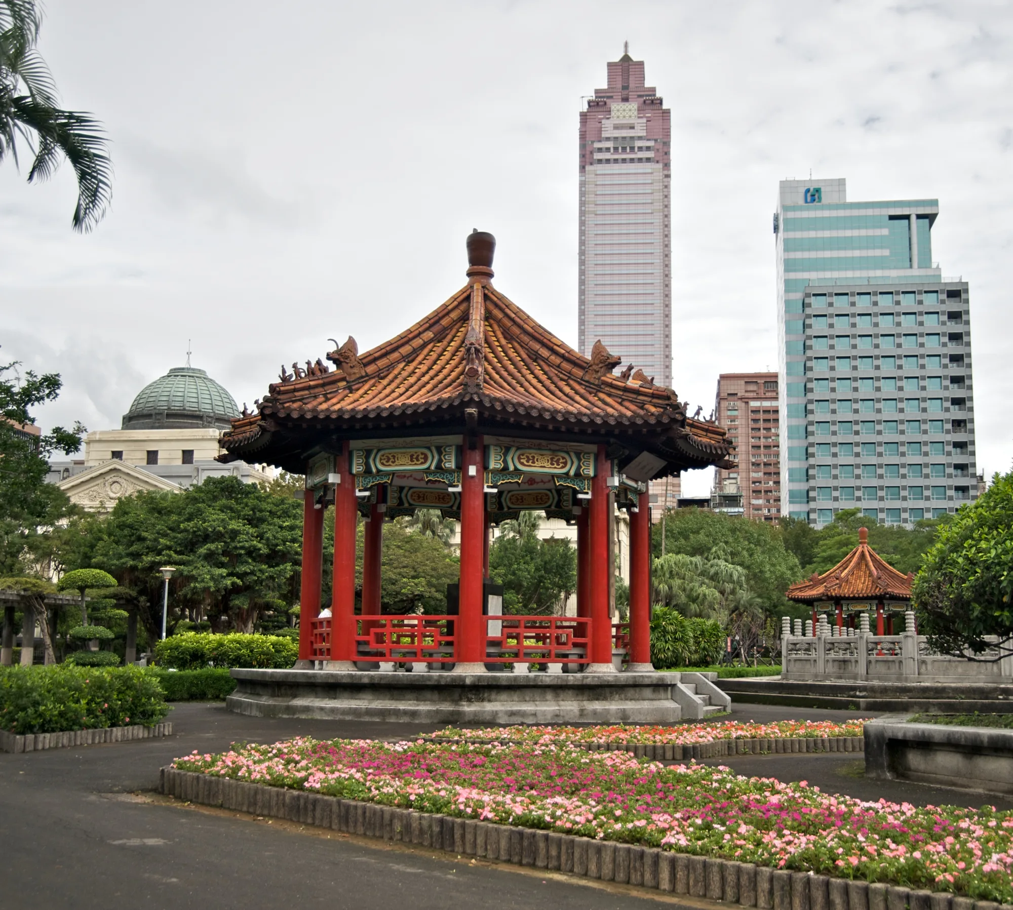 The red-pillared Chinese pavilion at 228 Peace Memorial Park in Taipei, with flower beds and Taipei high-rises behind