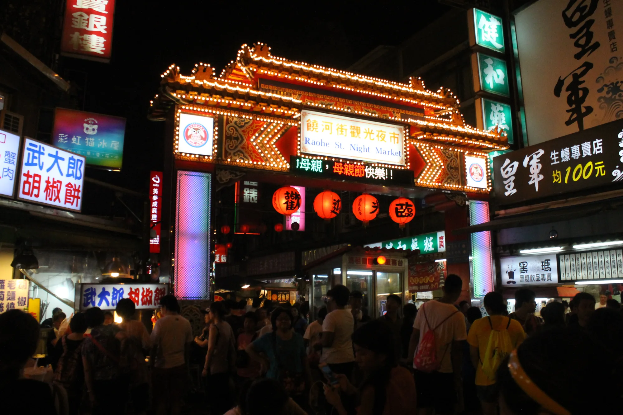 The illuminated traditional entrance gate of Raohe Street Night Market in Taipei with red lanterns and a dense crowd