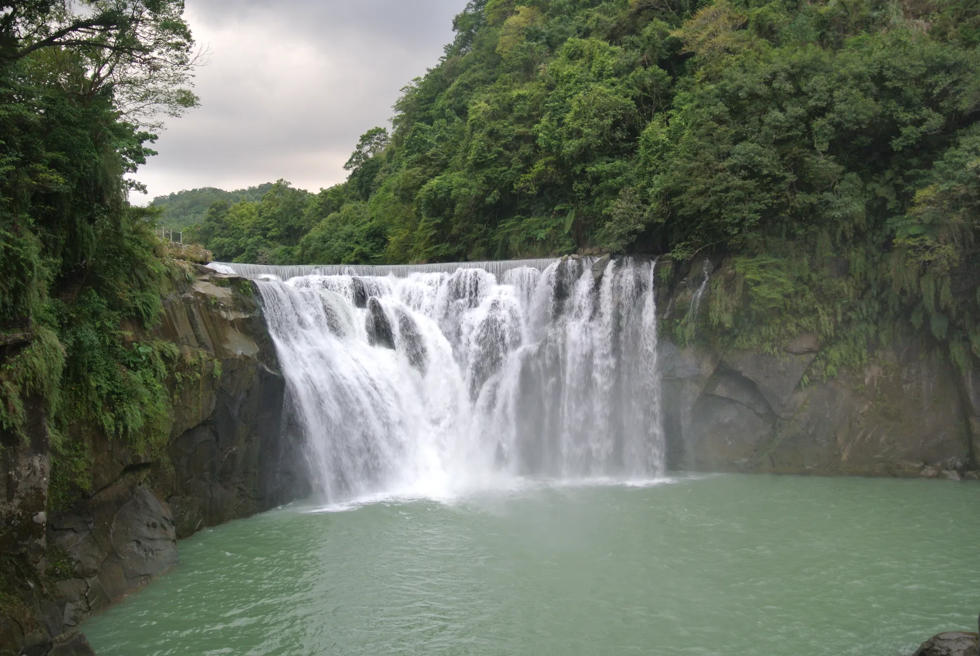 The wide curtain of Shifen Waterfall cascading into a turquoise plunge pool, framed by green forest, in Pingxi, Taiwan