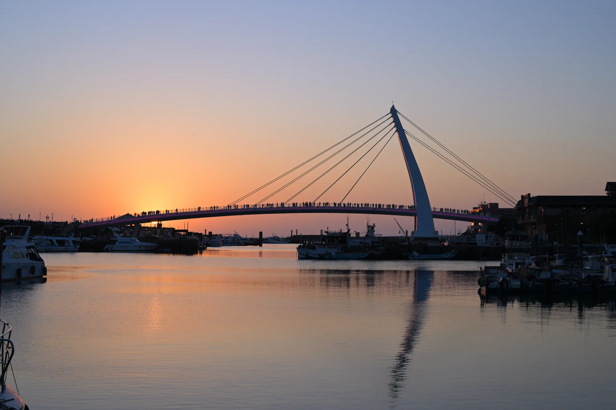 Tamsui Fisherman's Wharf Lover's Bridge silhouetted against a glowing orange sunset with boats moored below